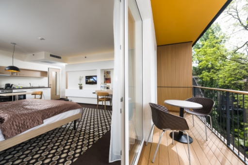 Modern hotel room with a double bed, patterned carpet, and a balcony featuring a small table and two chairs overlooking green trees.